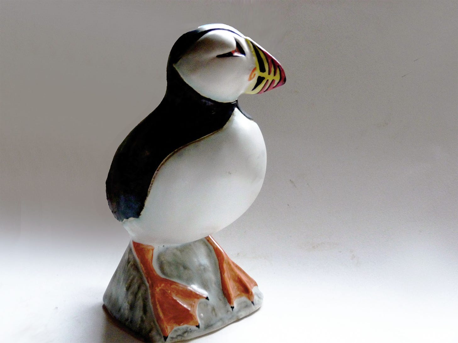 A stoneware slip-cast Puffin designed by Weston Neil Andersen with his affinity for the geometry of natural forms. The back and the mask-covered face is glazed in glossy ebony. The Full ripe chest is glazed in white matte, accentuated by bright coral wide webbed feet. The large dramatic beak is decorated in bright red, yellow, and black overglaze colors in a curved stripe pattern.