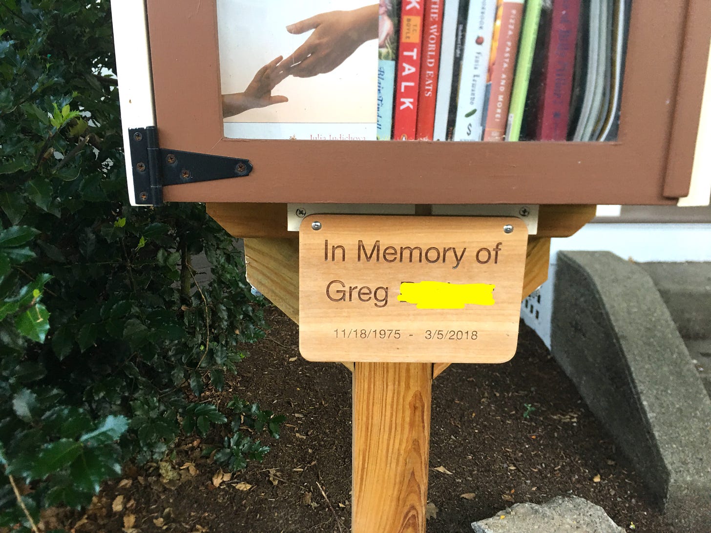 A Little Free Library made of wood features two jammed shelves of books, and a sign underneath that reads: In Memory of Greg, with his birth and death dates.