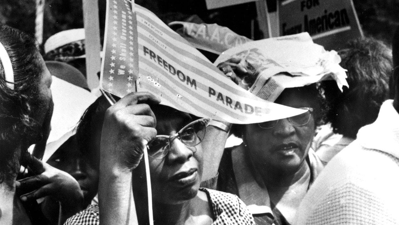 A black and white still of a Black woman holding up a pennant that reads Freedom Parade