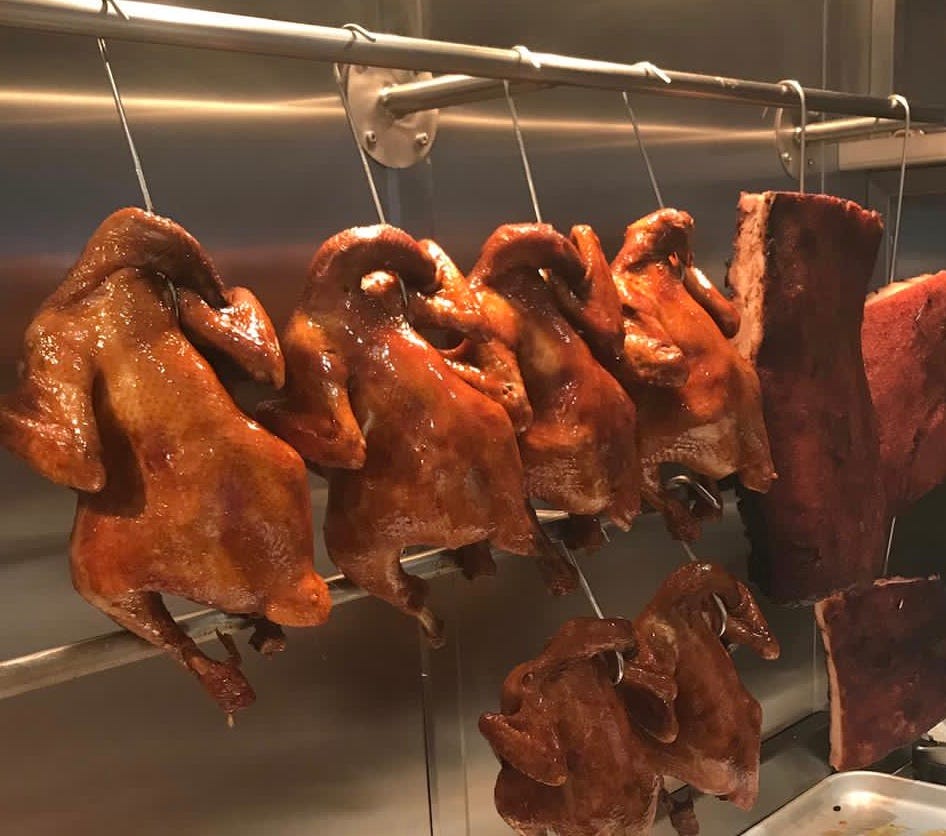 A row of four deeply browned cooked chickens hanging on hooks under a lamp in at a station in a professional kitchen, surrounded by stainless steel