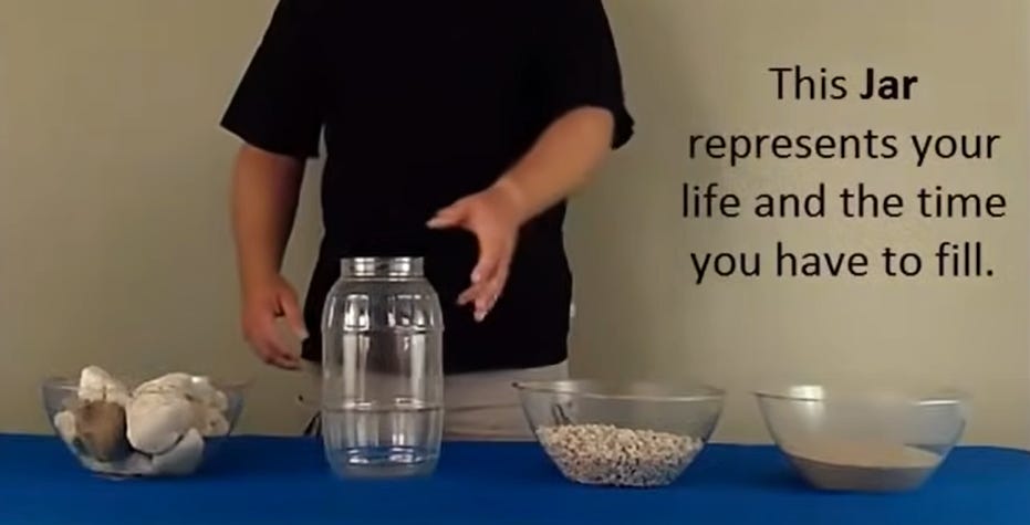 Screenshot of the video. Four things are on the table: a bowl of rocks, an empty jar, a bowl of pebbles, and a bowl of sand. A person is standing behind the table. There's a text that says: "This jar represents the life and the time you have to fill."