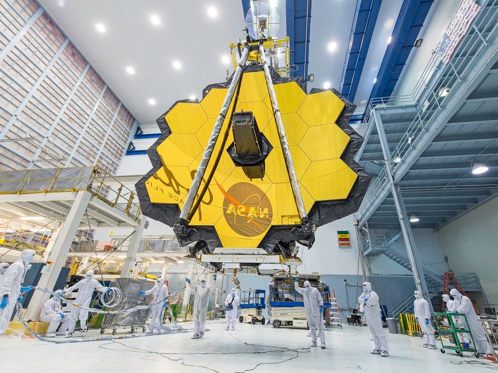 An image of the James Web Telescope in a NASA laboratory. Technicans are seen standing under it while the honey come shaped telecope mirror hangs above. The NASA logo is seen reflected in the mirrors.