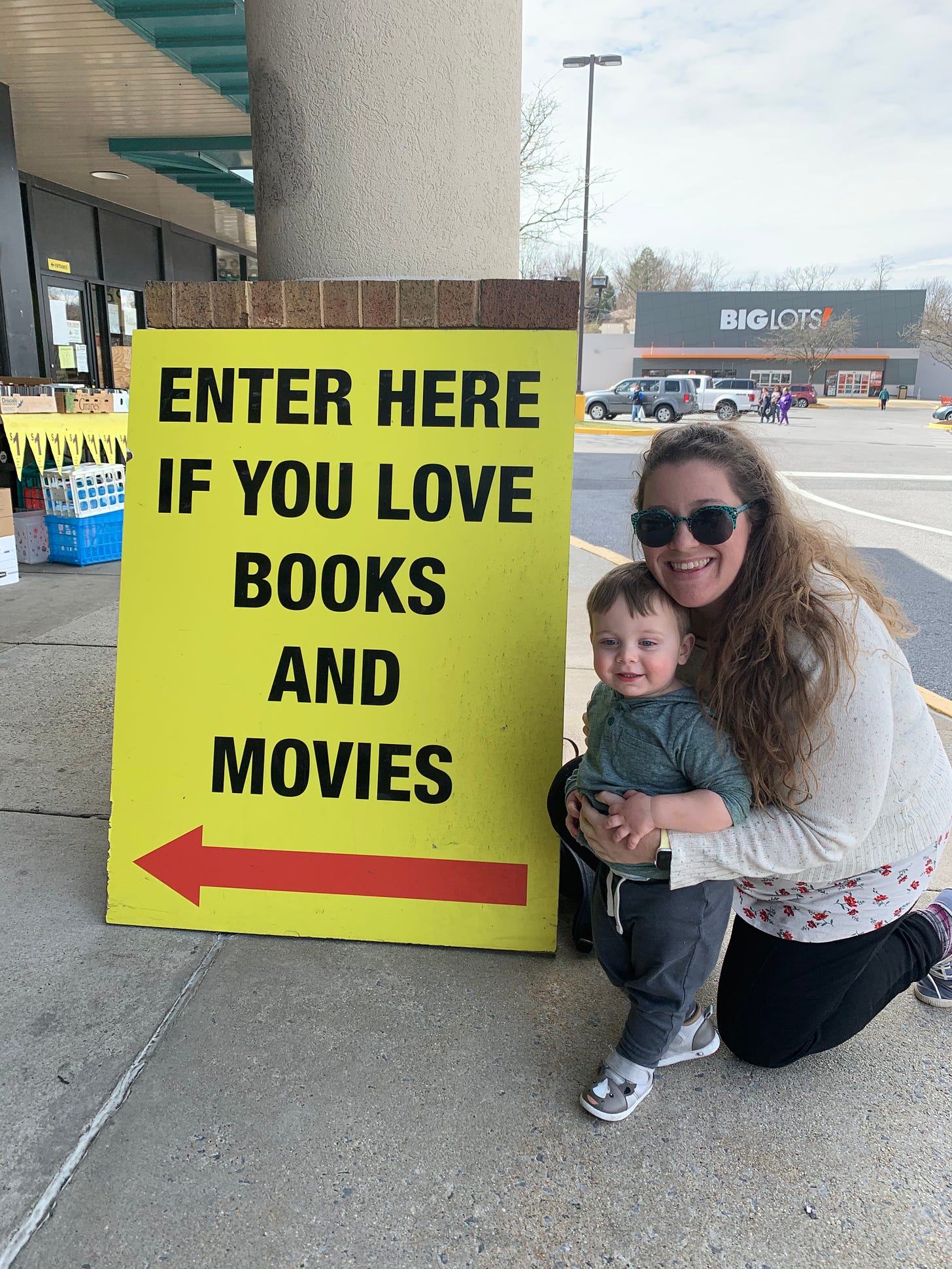 Marissa and her young godson pose, smiling, in front of a large, yellow sign that reads in black letters, "enter here if you love books and movies" in a Maryland strip mall.
