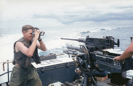 On a boat with waves in the background, a young man with blonde hair holds a video camera to his face. He wears a military vest and pants with bare arms. In front of him--the subject he is filming--is a gun or other military equipment on the boat. An operator's forearm is visible holding/manipulating the gun or equipment. 