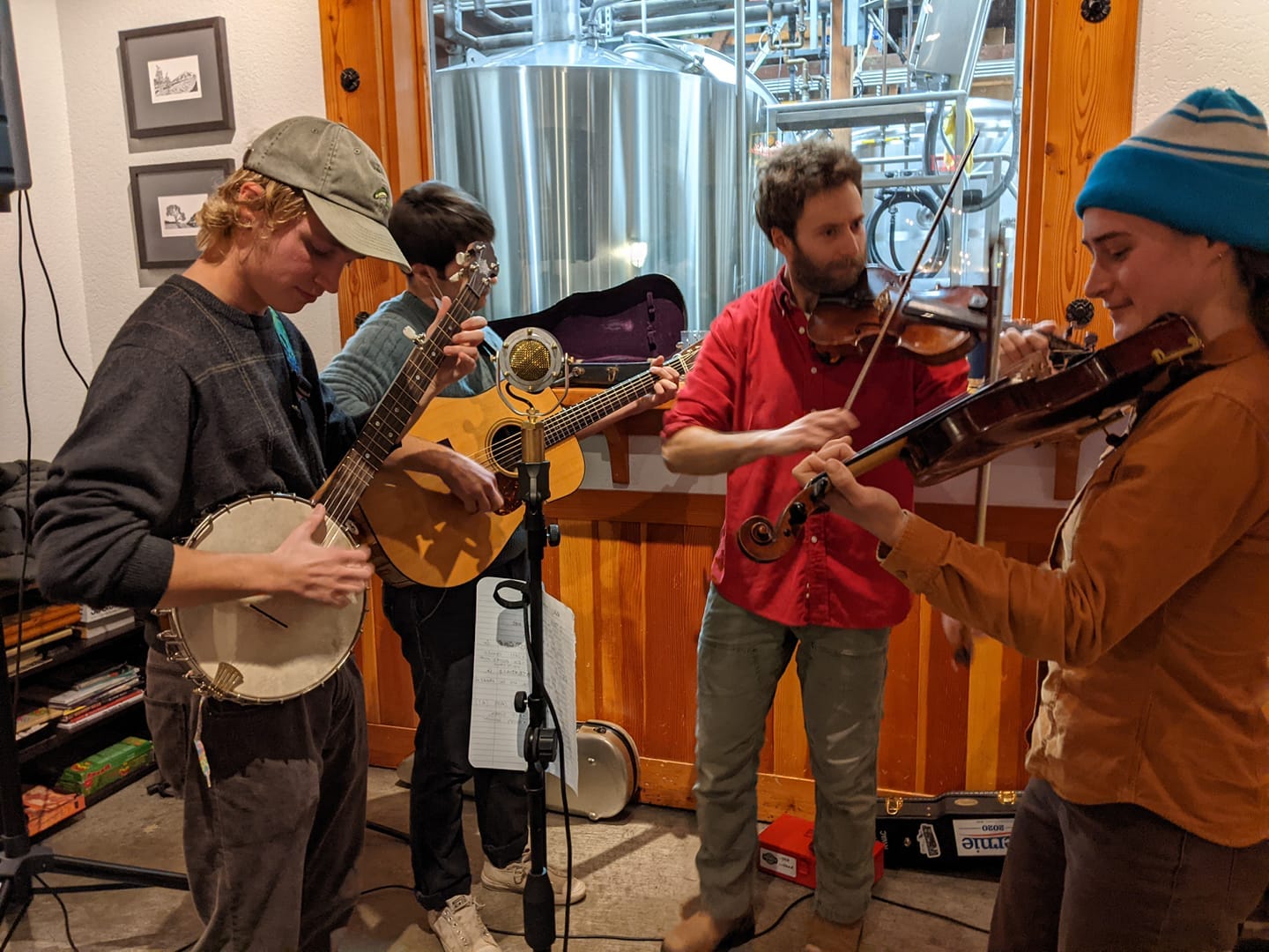 A guitarist, a banjo player, and two fiddlers, with brewing equipment in the background
