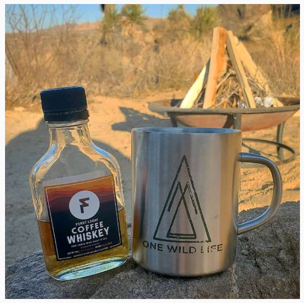 A mini-bottle of First Light Whiskey and a silver metal coffee mug with the One Wild Life logo sit on a rock in the foreground. In the background a sunset lit metal campfire ring filled with wood to be lit for a campfire is blurred out along with sand and Joshua Trees in the desert.