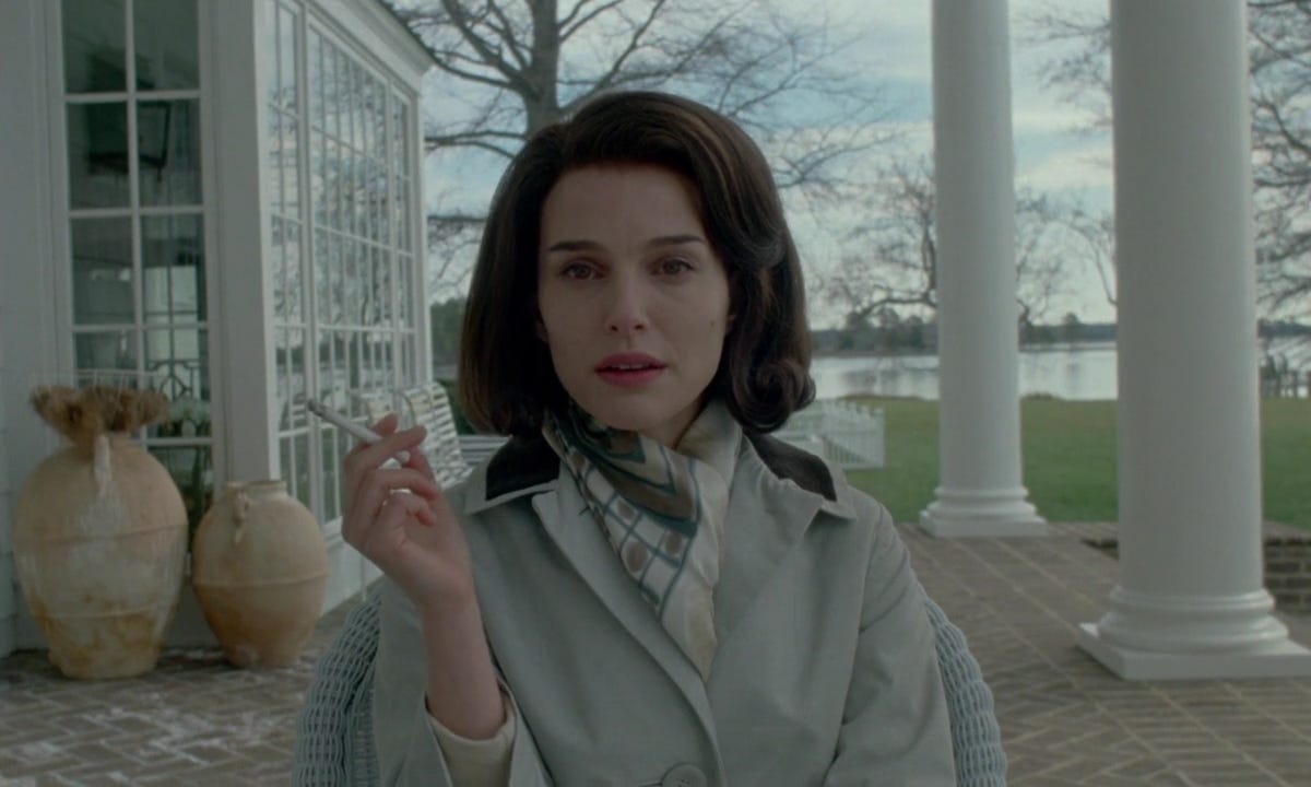 Natalie Portman as Jacqueline Kennedy Onassis in Pablo Larraín’s Jackie (2016).  She is smoking on a verandah in a tan jacket and white scarf. 