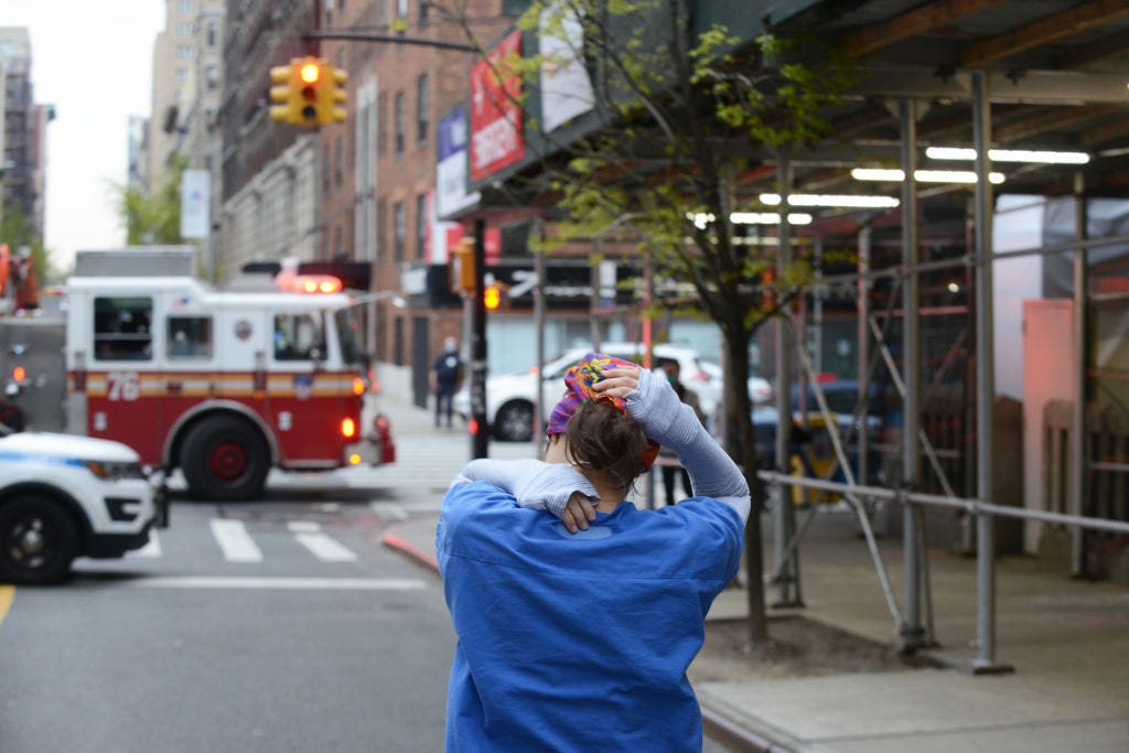 A nurse in scrubs stands on the streets of New York, which have been blocked by a fire truck, facing away from the camera and holding her head and shoulder in her hands.