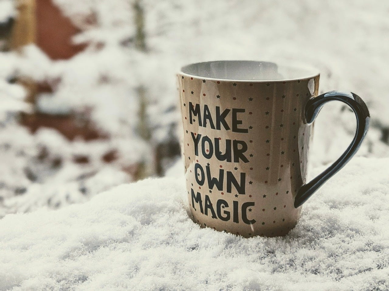 Mug on snow covered outdoor windowsill there is a blurred background of snow covered trees. The mug’s text reads ‘make your own magic’