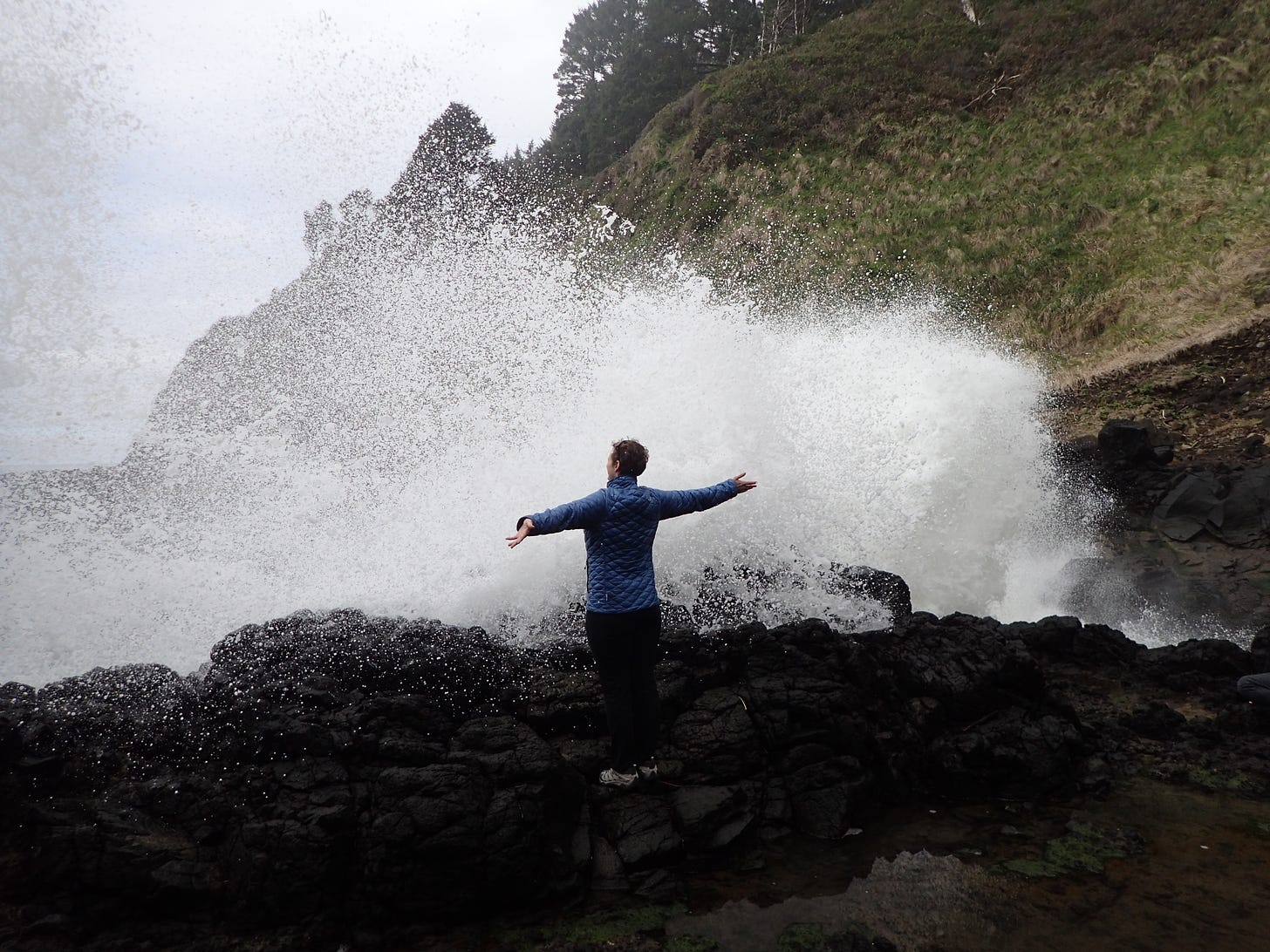a woman on a rocky outcropping with an incoming wave and her arms stretched out to meet it
