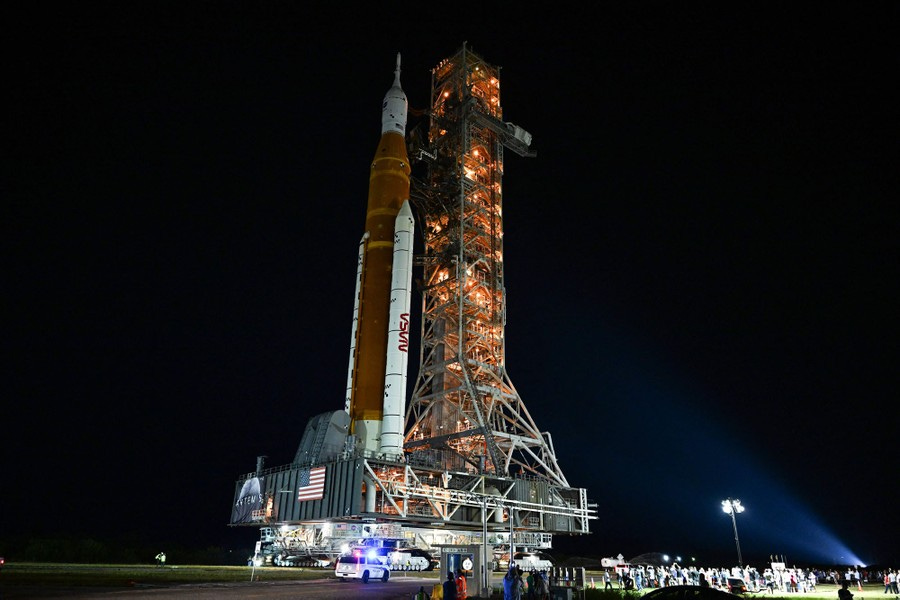 A tall rocket ship stands on a mobile platform beside a tall tower, lit up at night.