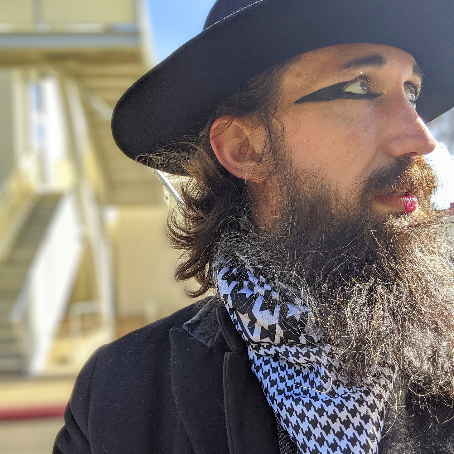 Jason Wyman looking to he right with the sun on their face. Jason is wearing a black wide-brimmed har with long brown hair and a long bushy beard. Jason has a houndstooth silk scarf on and a black wool coat. Jason is wearing bold black eyeliner and a pink lip. Jason is set against a blurred out background of a yellow home.