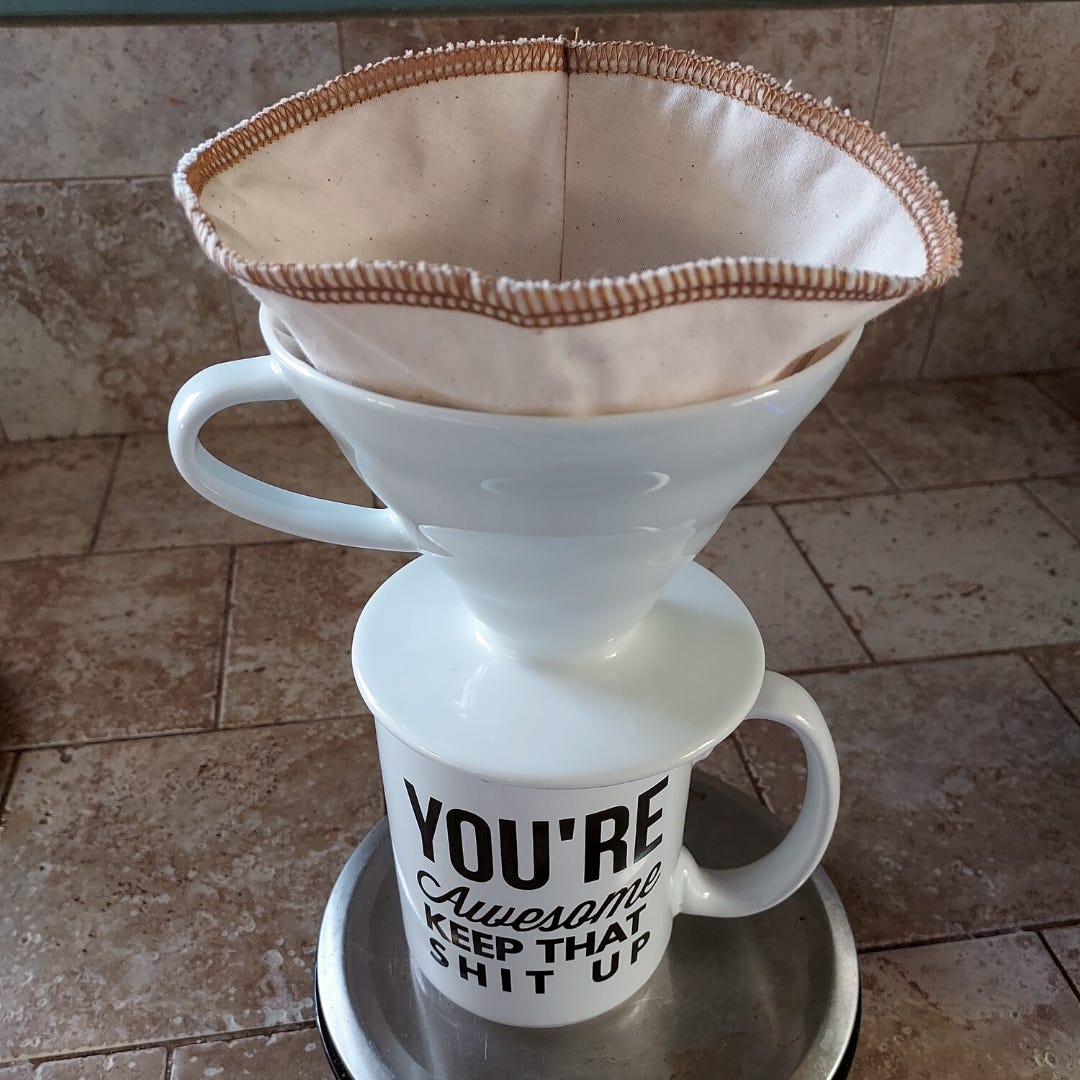 A cloth filter in a V60 pour over coffee brewer on top of a white coffee mug sitting on a beige tile kitchen counter-top from above.