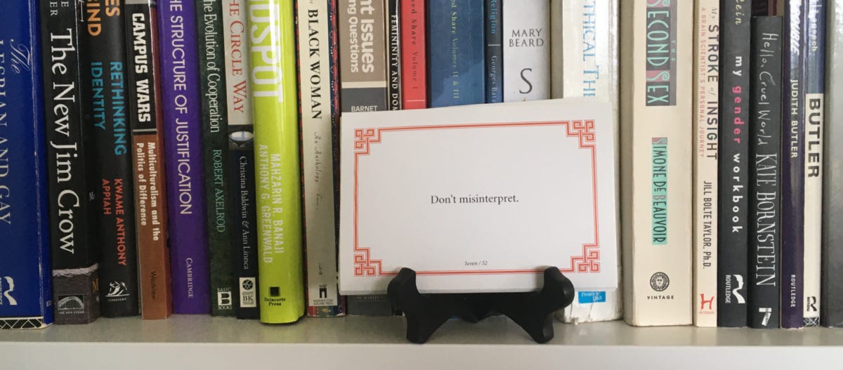 Lojong slogan card on a black card stand on a white bookshelf with books behind it. Card reads: Don’t misinterpret