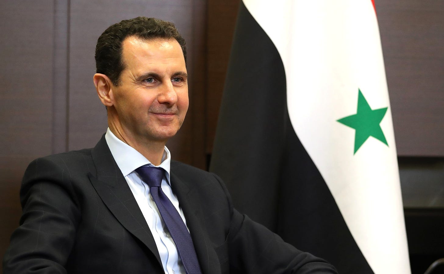 A man grins with a sneer in front of a Syrian flag.