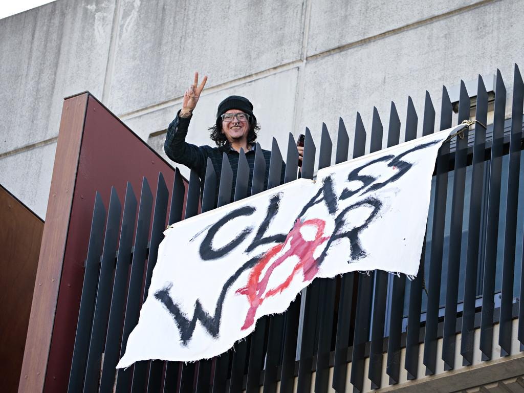A young anarchist smiles down from a balcony, holding up two fingers in a V above a banner that reads “CLASS WAR” with the A for anarchy symbol.