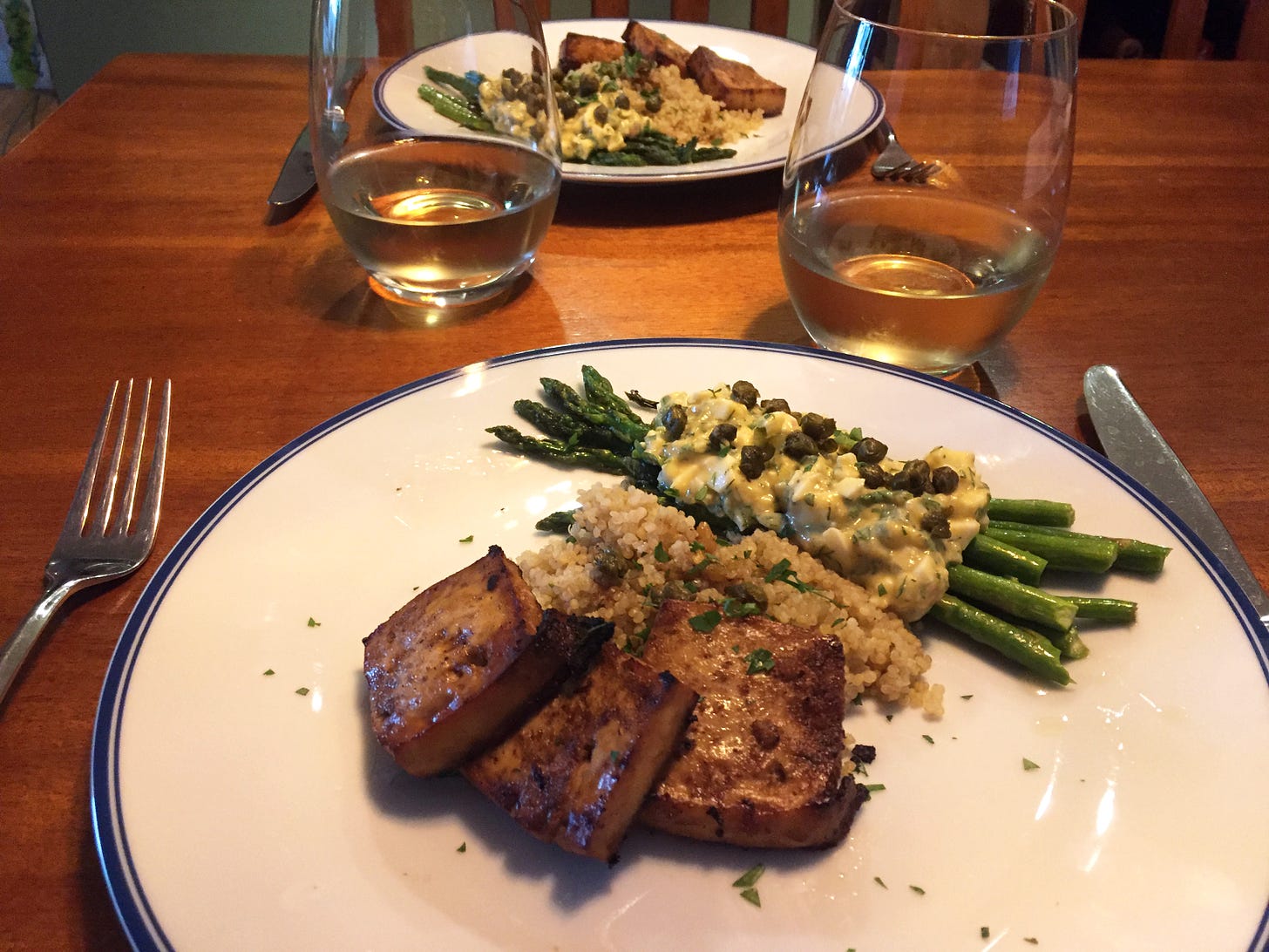 Two plates across from each other, each with browned marinated tofu arranged over quinoa with asparagus gribiche and capers. Stemless glasses of white wine sit besides the plates.