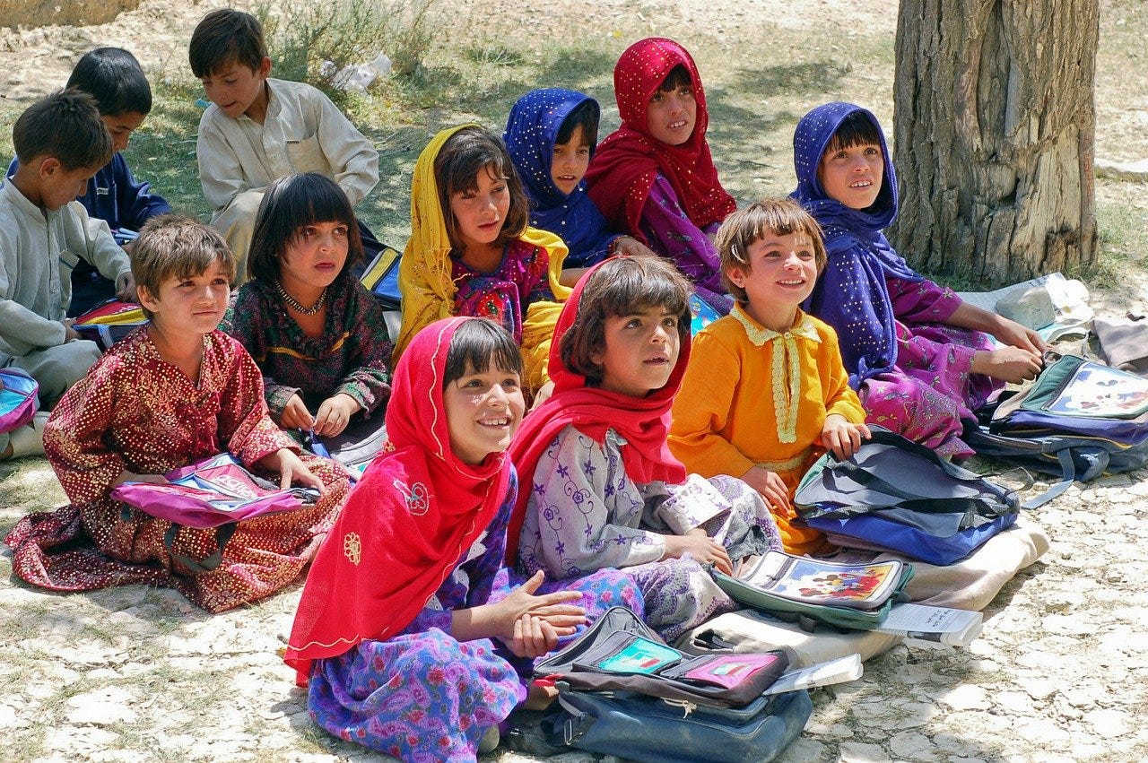 Muslim children in colorful scarves, all sitting outside on the ground for class.