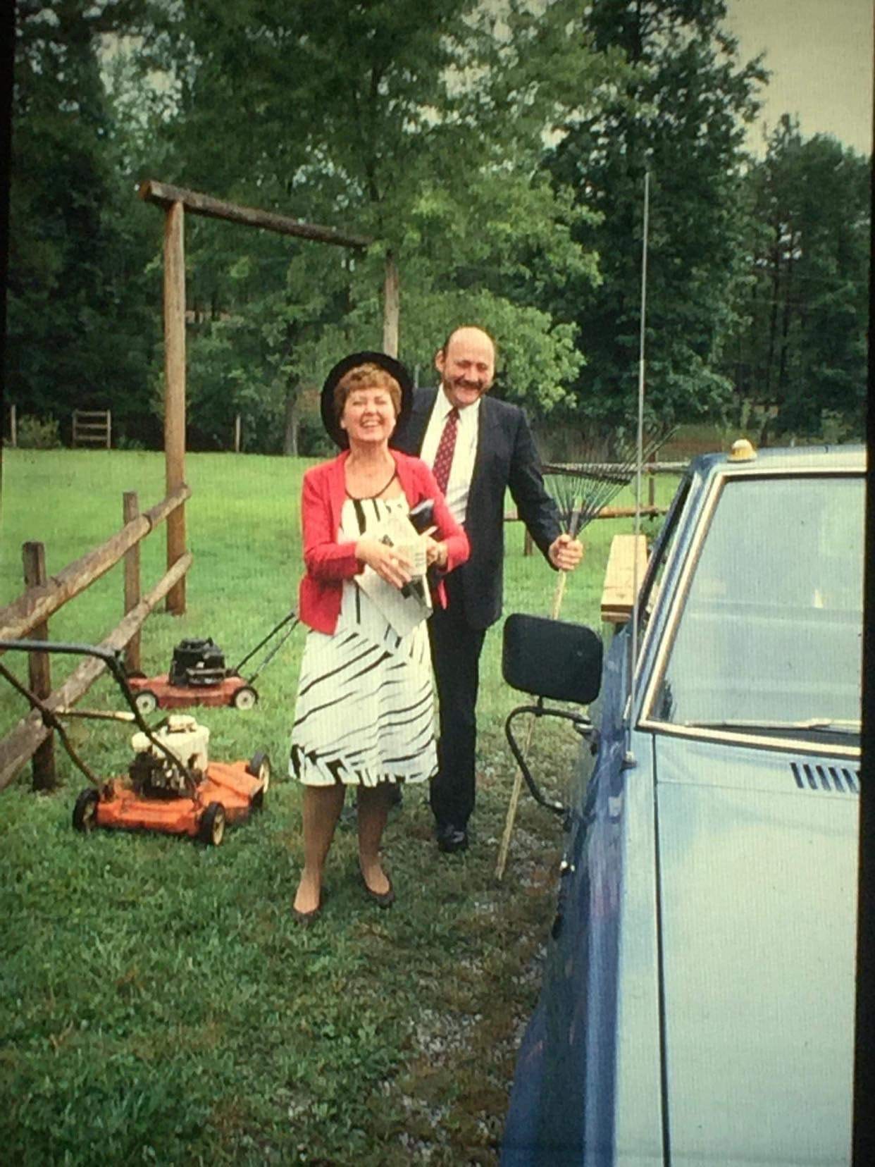 This picture was probably taken in the '80s or '90s. It's of the author's maternal grandparents and they are featured in front of a wooden post fence in an expansive property with trees, ca blue car, and two lawn mowers scattered around them. They are in the center of the photo, my grandmother wearing a black and white dress and a pink jacket and a black hate and holding a newspaper and purse while my grandfather is behind her in a suit, red tie, holding a rake upside down. They look a bit like American Gothic. They probably just got home from church.