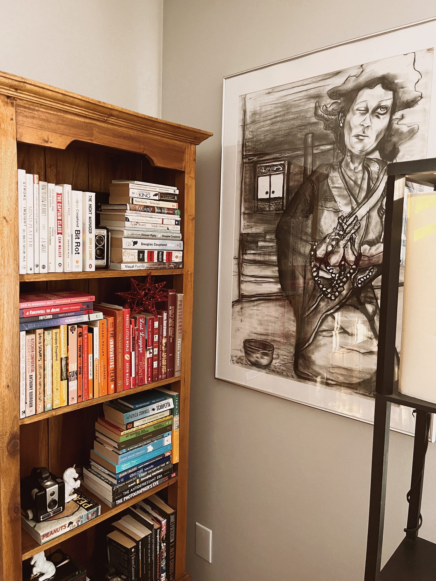 Large charcoal drawing of a woman clutching prayer beads and looking askance at the heavens, one eye open. Next to the picture is a bookshelf where I've organized my books by colour of the covers.