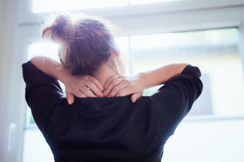 Rear view of a young woman looking out the window as she massages her neck.