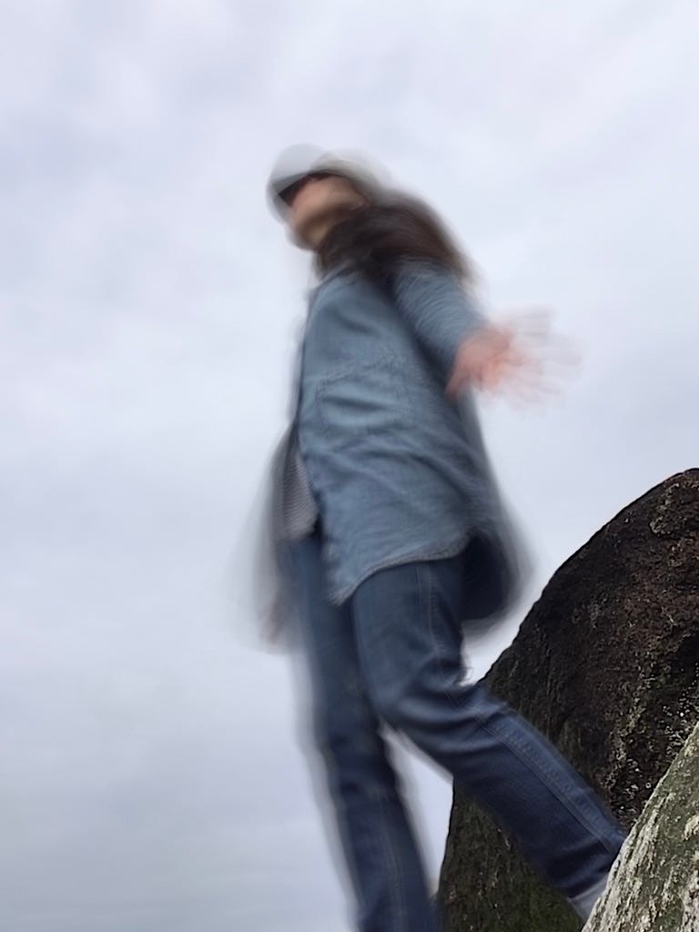 long exposure fuzz/blur of Cassandra, white two-legged, two-armed human woman standing on her feet with arms outstretched at her sides (only her left can be seen) she is facing the left, the camera is on the ground looking up at her. there are two boulders behind her that are somewhat covered in barnacles. the sky is gray-ish behind her