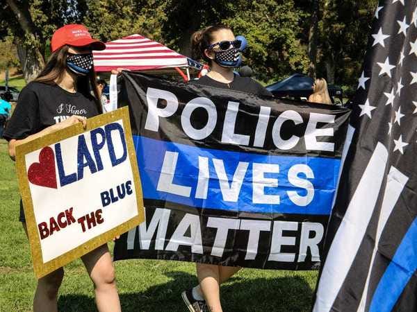 Two white "Back the Blue Protesters" carry signs. One reads "Police Lives Matter." The "thin blue line" bastardization of the U.S. flag can be seen.