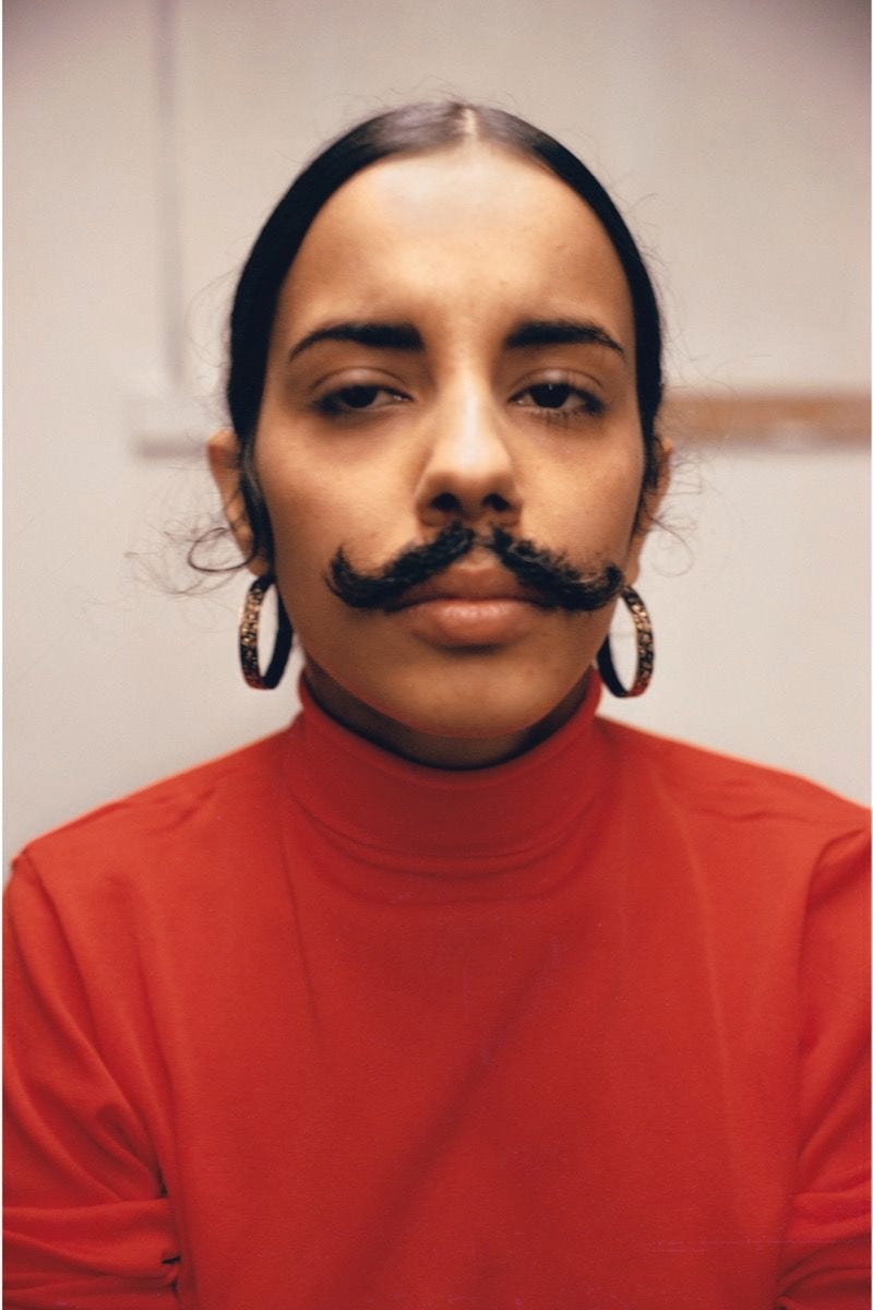 The artist in a red turtle neck sweater with her hair tied back in a bun. She's wearing shiny hoop earrings, and has a mustache taped above her lip. She is staring into the camera