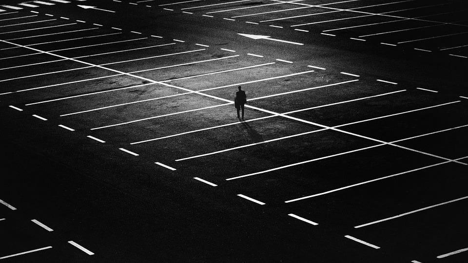 A silhouetted man alone in the middle of a large, empty parking lot at night