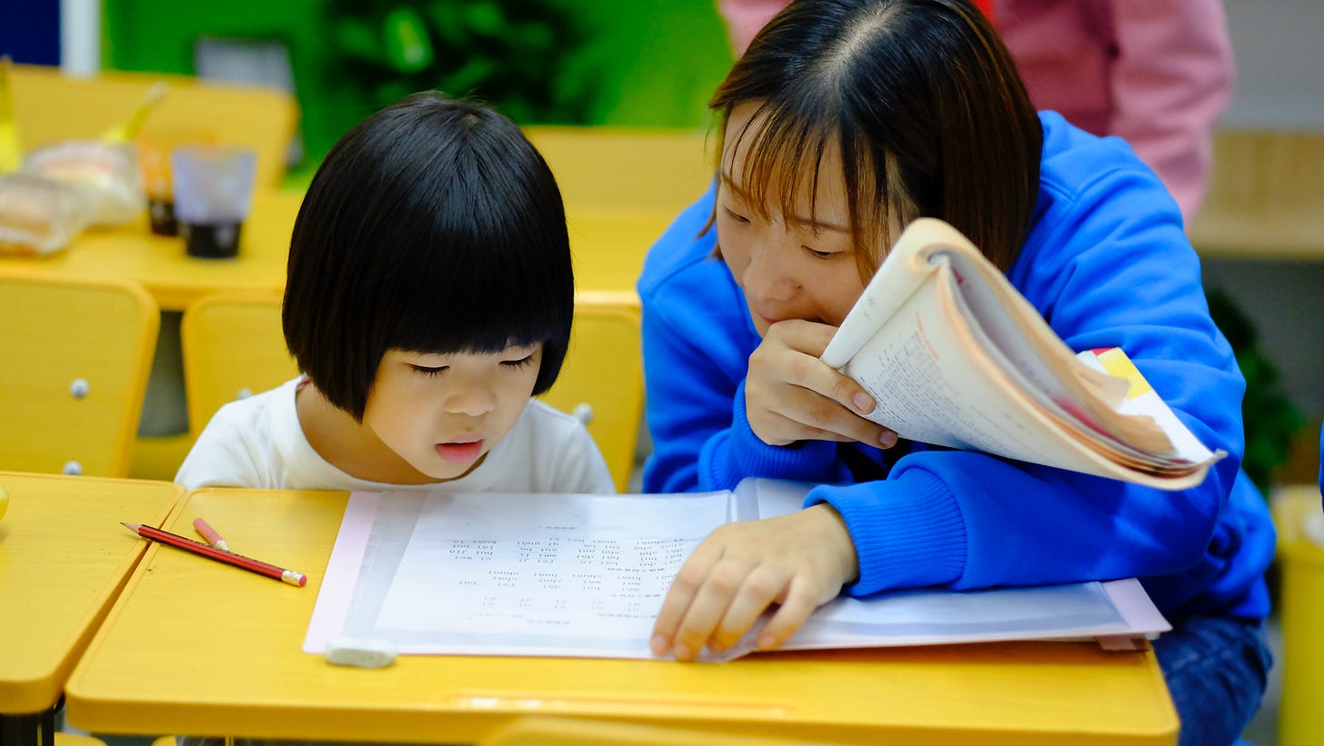 Chinese girl sitting at desk looking at paper with her teacher