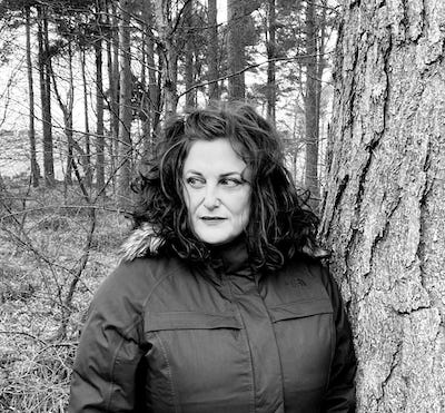 A black and white photo of a woman in a forest leaning against a tree. She is dressed in a parka and is looking at something off to her right. She is Allyson Shaw