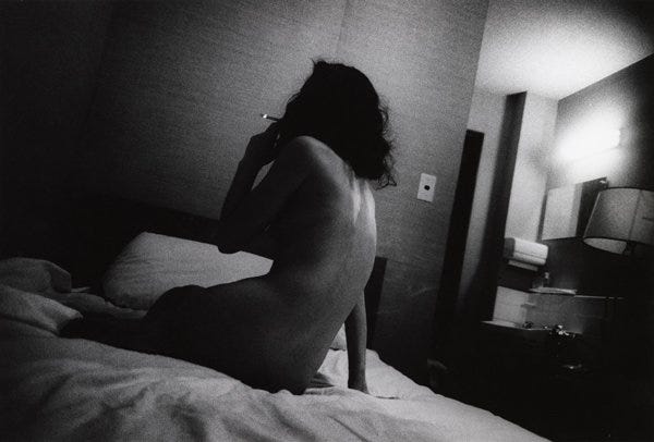 Fotografia em preto e branco de uma mulher nua, sentada na cama, de costas para o fotógrafo. Ela tem uma mão apoiada no colchão e com a outra fuma um cigarro. Seu rosto está oculto pelas sombras do cabelo, muito preto, cortado na altura dos ombros.