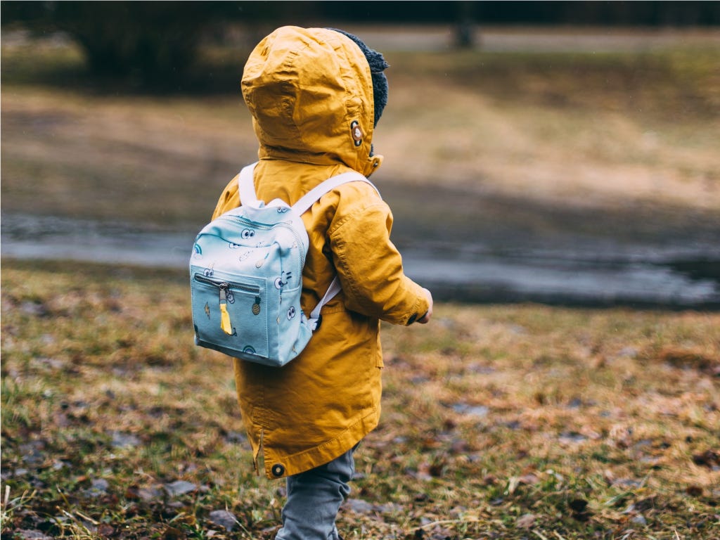 A young child in a yellow coat and a backpack stands with their back to the camera. Photo by Daiga Ellaby on Unsplash