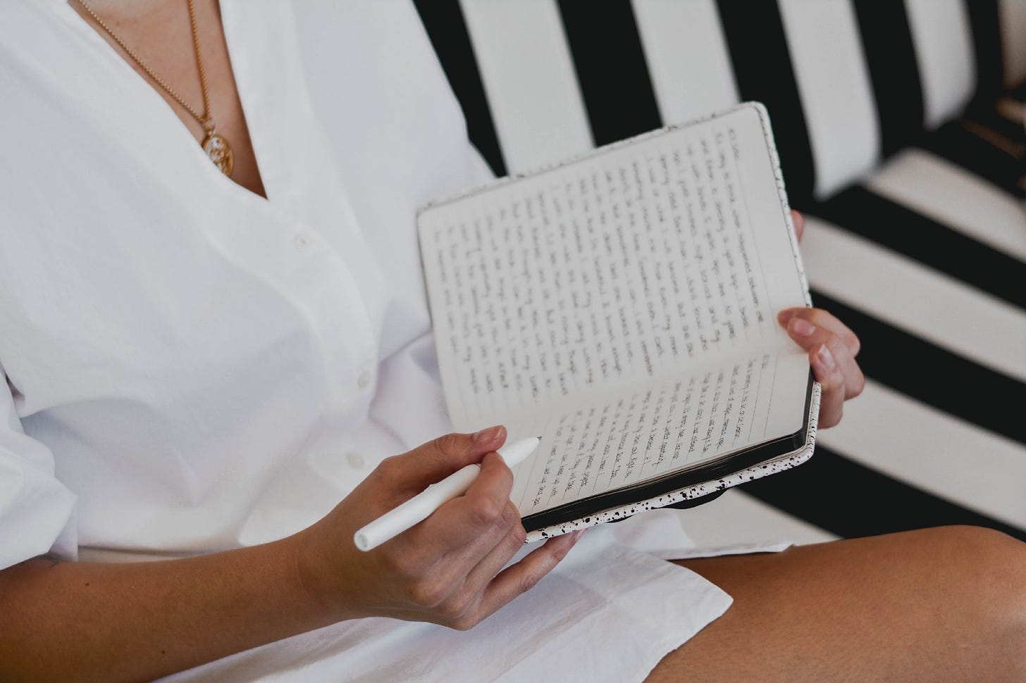 Woman sitting on striped couch with a baggy white button up shirt on, journaling her morning pages. A concept created by Julia Cameron, author of the Artist’s Way.