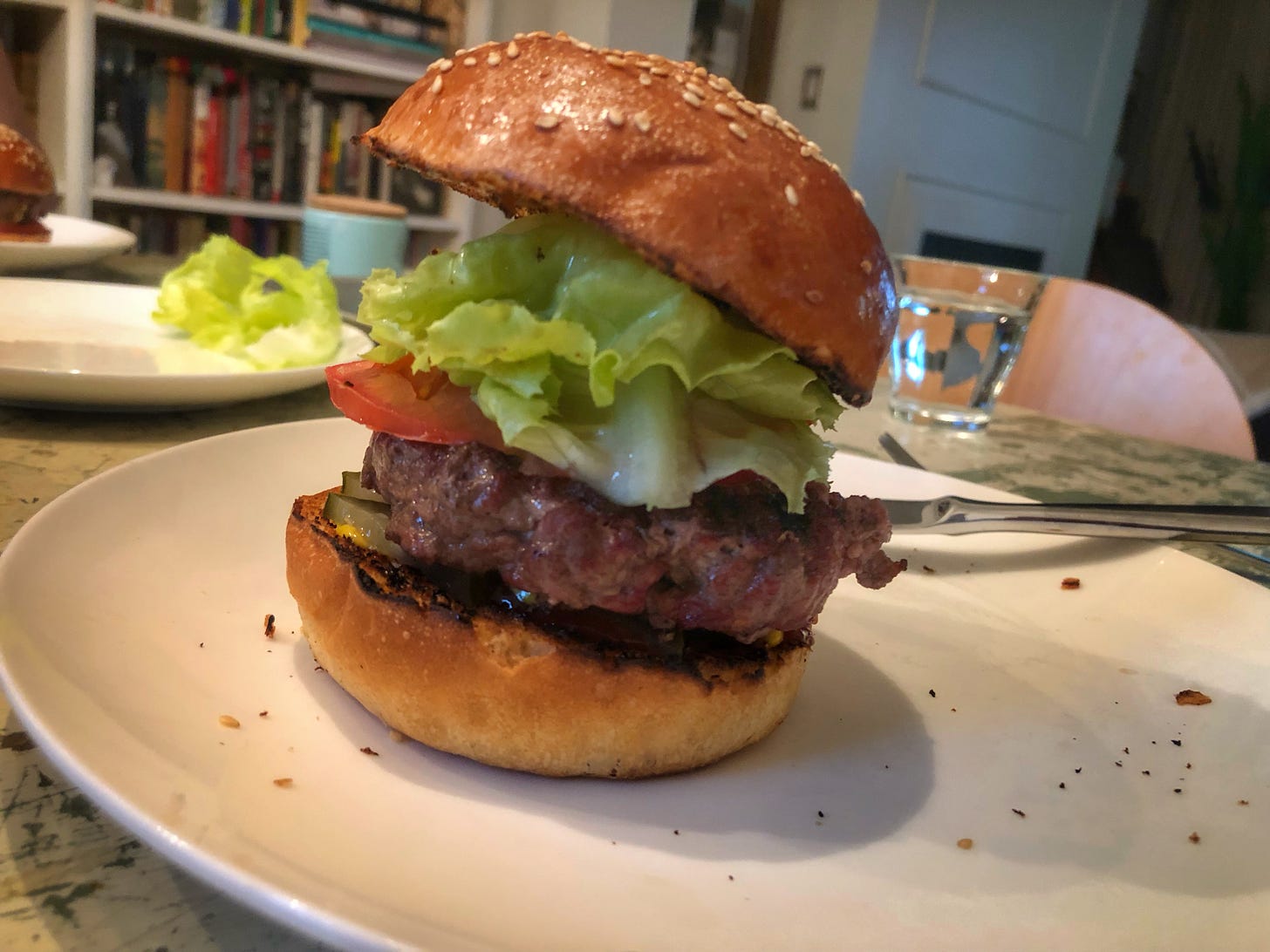 A dinner plate with a burger on a sesame seed bun, piled high with lettuce and tomato. It is far too tall to fit into someone's mouth.