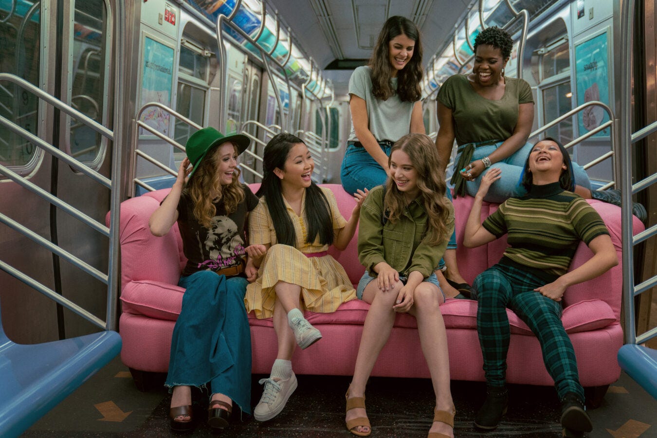 Still from To All the Boys: Always and Forever of the main character and her friends sitting on a pink couch on the subway. Two of them are sitting on the back and four are sitting on the couch.