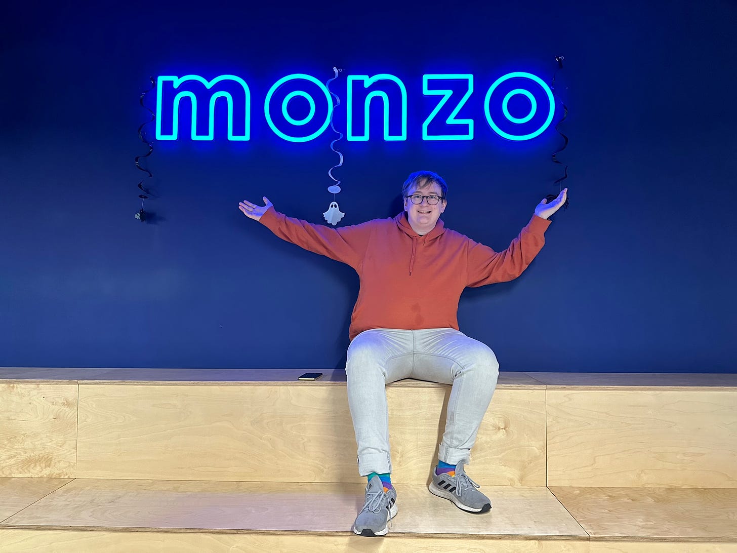 Avery is sitting down in an orange hoodie and light blue jeans, arms outstreached beneath a large neon Monzo logo. They're smiling to the camera.
