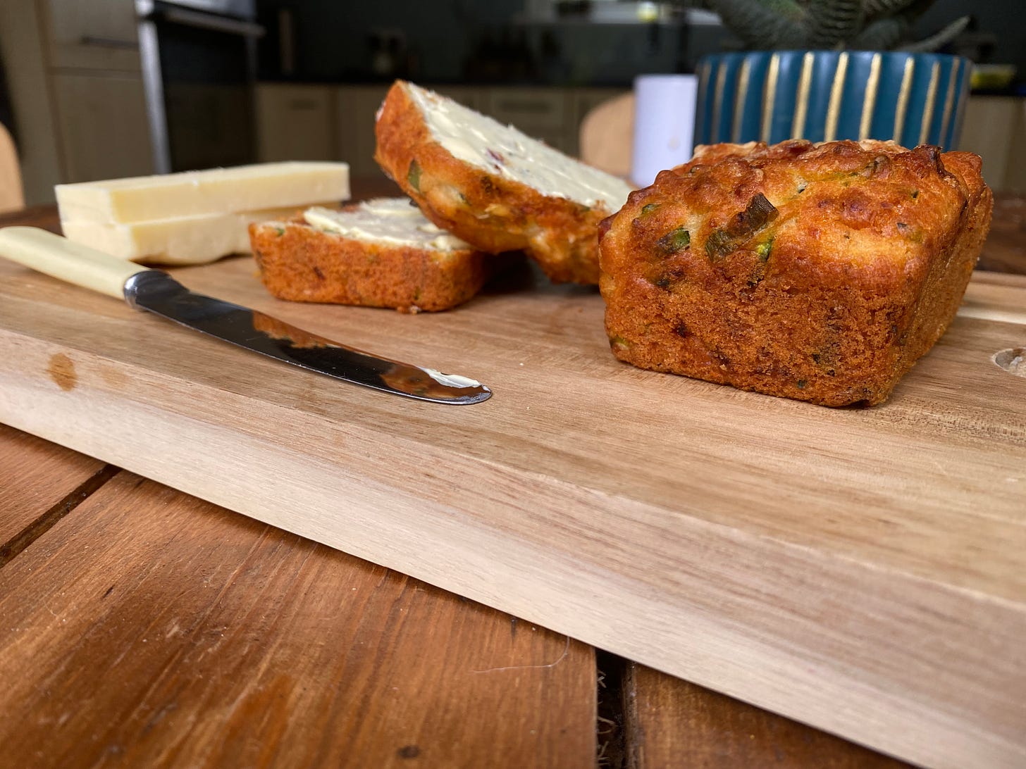 Courgette mini loaves on a chopping board next to two small wedges of cheese. A knife with butter is also on the wooden board. 