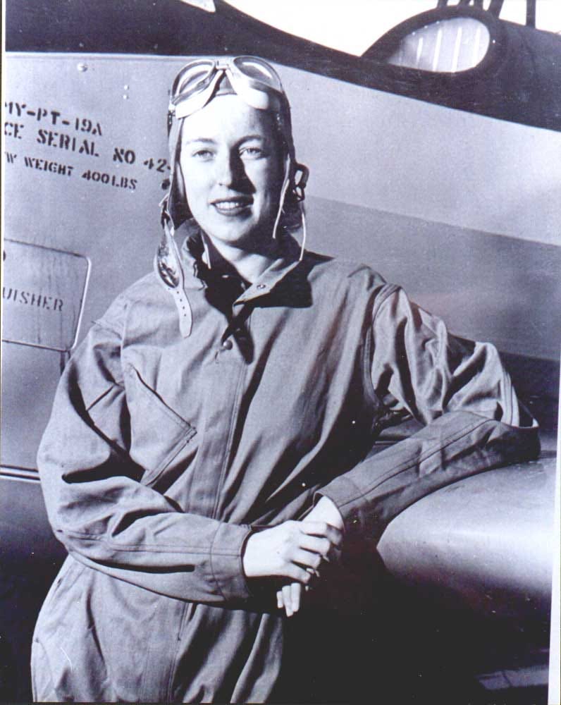 Cornelia Fort, in flight gear, leans with one elbow on the wing of the plane and smiles at the camera.