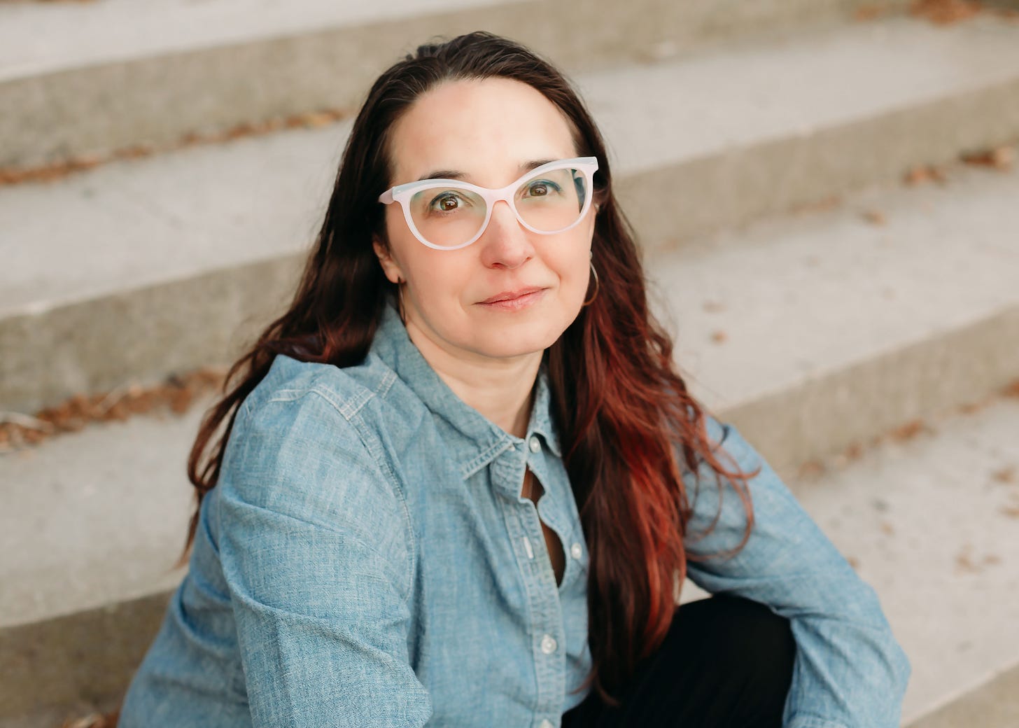 Headshot of author Sonya Huber from the waist up, wearing a denim button-up and white-rimmed glasses, sitting on concrete steps.