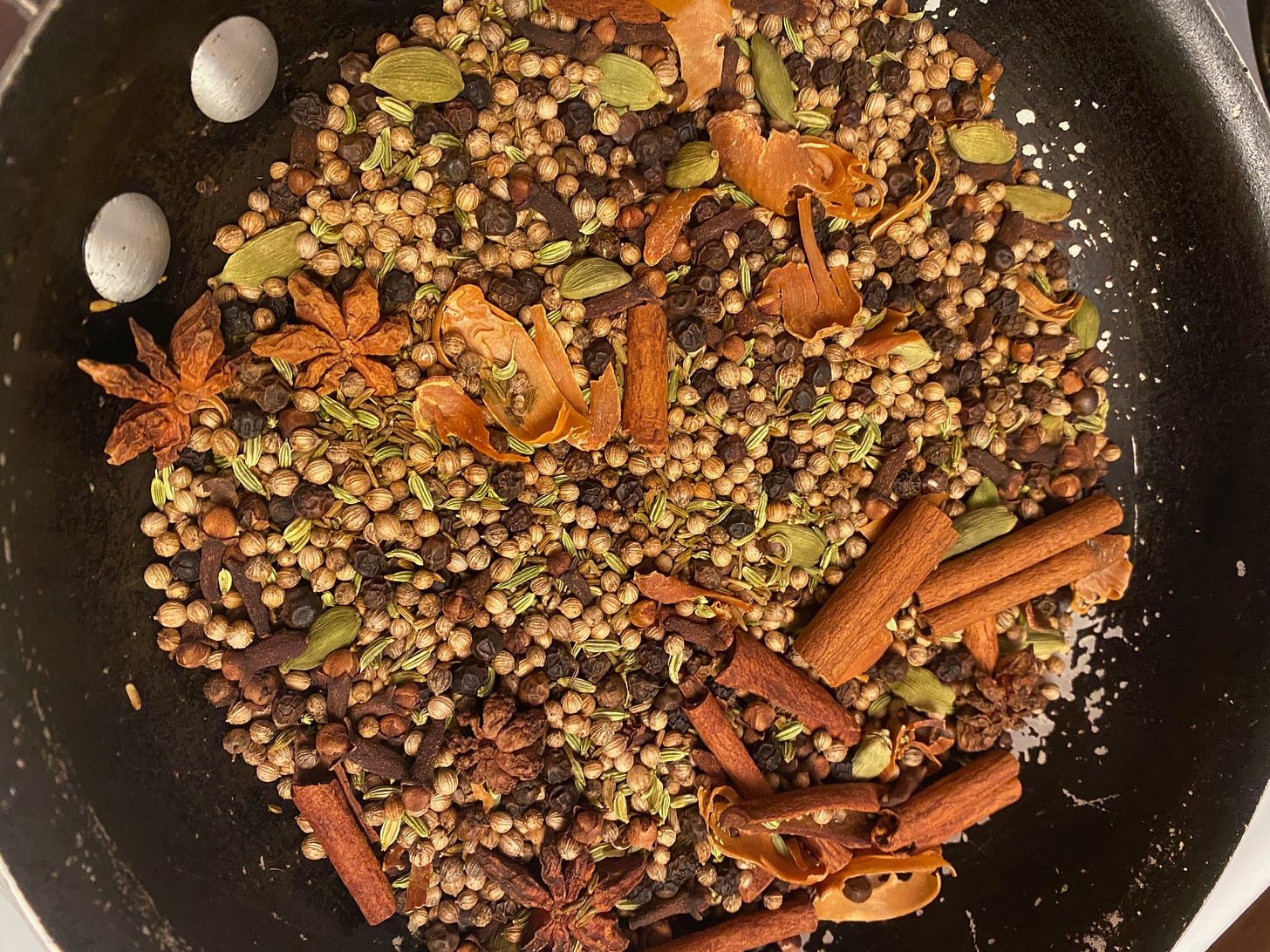 An array of whole spices in a small frying pan—green cardamon pods, star anise, broken cinnamon sticks, cumin, coriander, cloves, fennel, and mace.