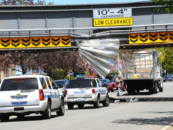 A busy road. Police cars are on the scene as a truck has struck an overpass whose low clearance is posted. The truck has had the roof and back door of its container car torn off, and much of the debris is still visible embedded into the structure of the overpass.