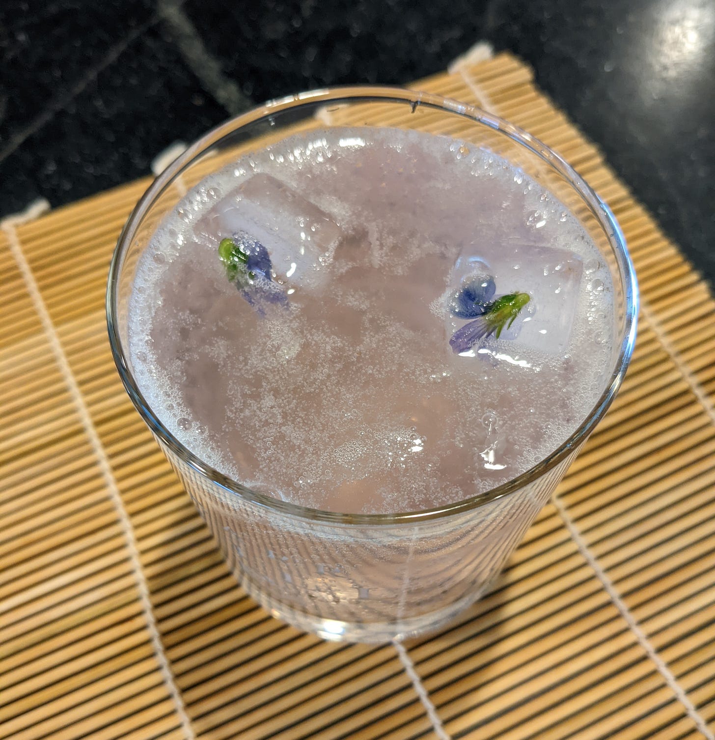 A small glass containing a fizzy lemonade drink that's just barely tinted purple. Two ice cubes with violets in them float in the glass.