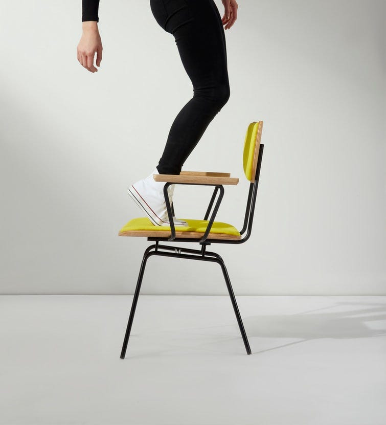 A photo of a yellow chair in the center of the frame against a black background. There is a person standing on the seat of the chair in converse and dark leggings, balancing on their toes. Only the lower half of the person's body is visible.