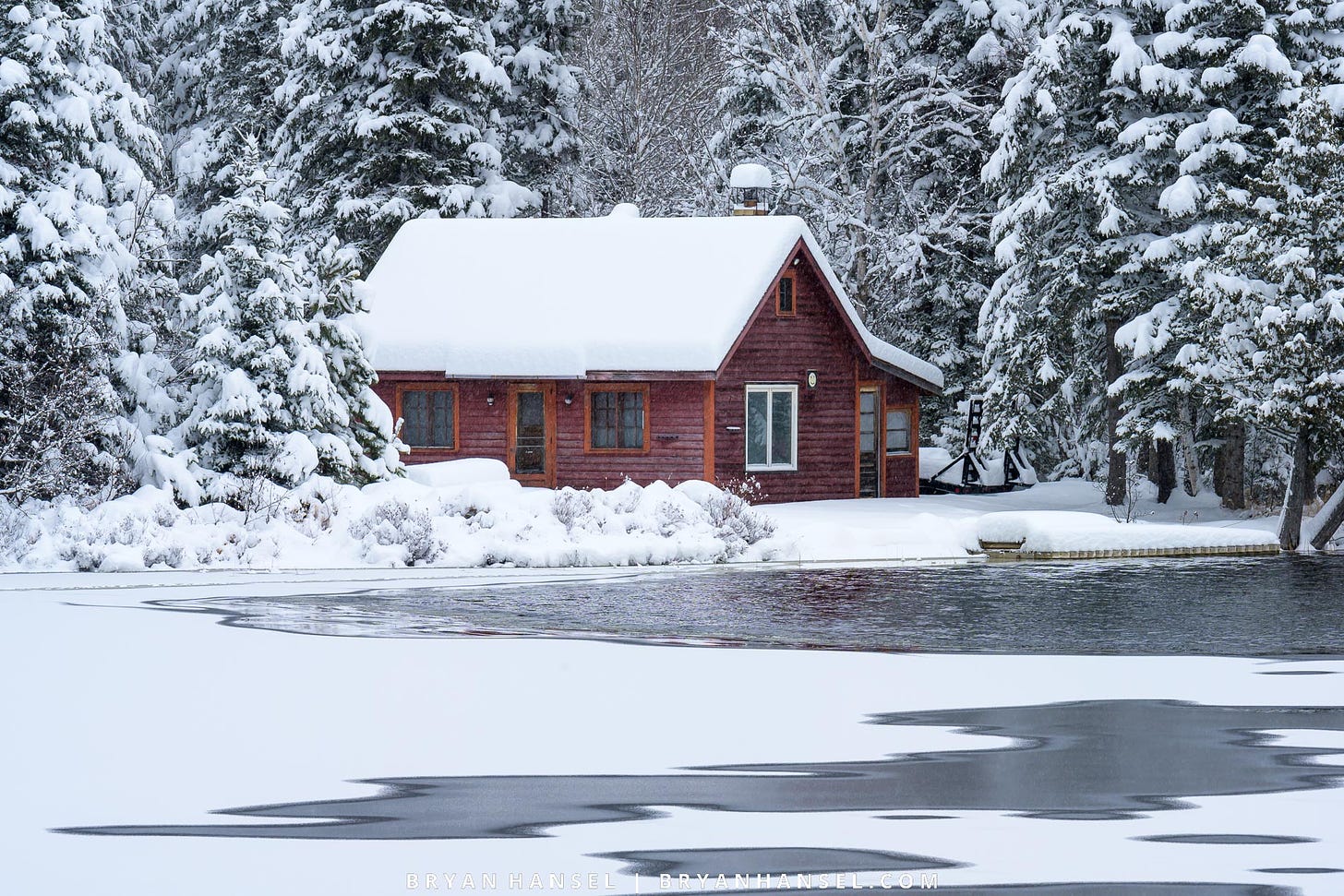 A photo of a red cabin covered in snow on a lake that is half frozen.