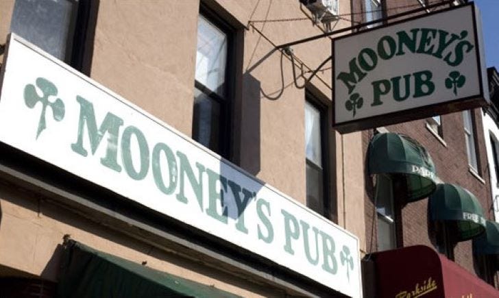 A daylight view looking up at two signs for "Mooney's Pub," green on white with stylized shamrock/crosses, hanging above the street.