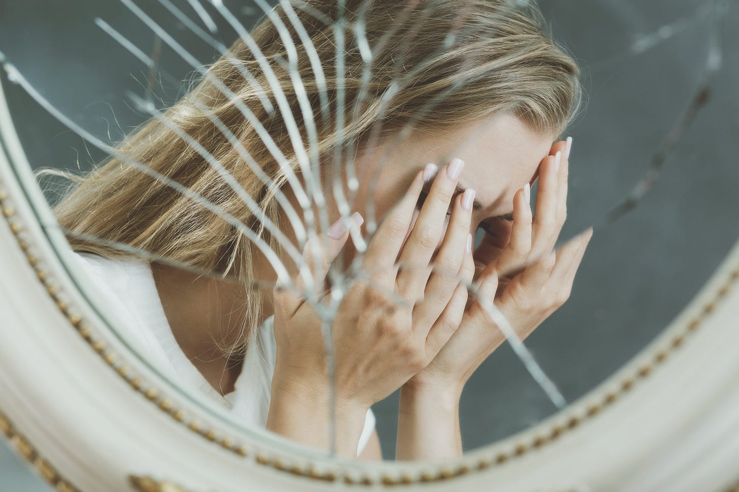 A woman holds her face in her hands and cries in the reflection of a shattered mirror.