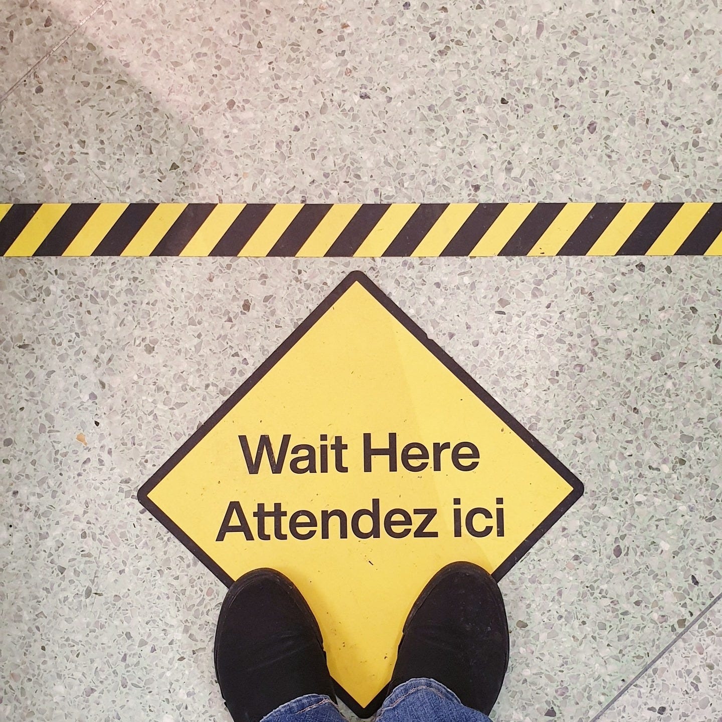 A shot of the ground with a yellow tape to make the start of the queue line and a yellow rhombus with the phrase "Wait here" in English and French.