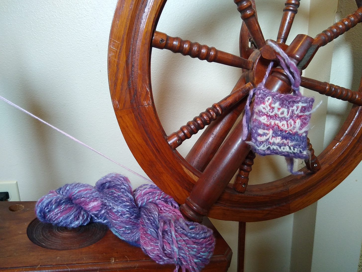 A skein of blue, purple, and pink yarn; a swatch that says "Start small. Then smaller." hanging from a spinning wheel.