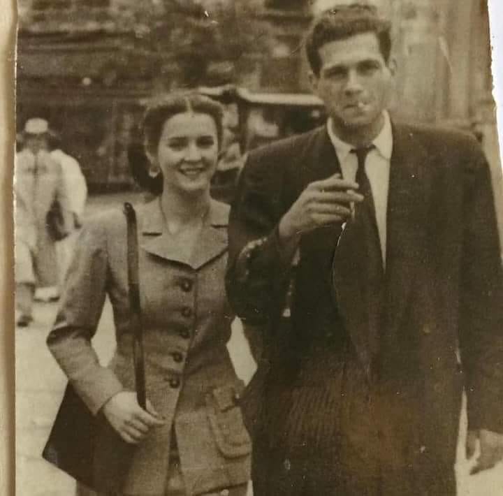 foto antiga, em preto e branco meio sépia, do hélio pellegrino fumando, de terno, e caminhando de braços dados com uma mulher de casaco e bolsa, os dois muito elegantes